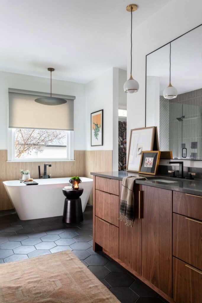 A bathroom with a freestanding white bathtub, a dark wood vanity with a black countertop, hexagonal black floor tiles, and pendant lights. The room has a minimalist decor with framed artwork, a small black side table, and a window with a beige roller shade.