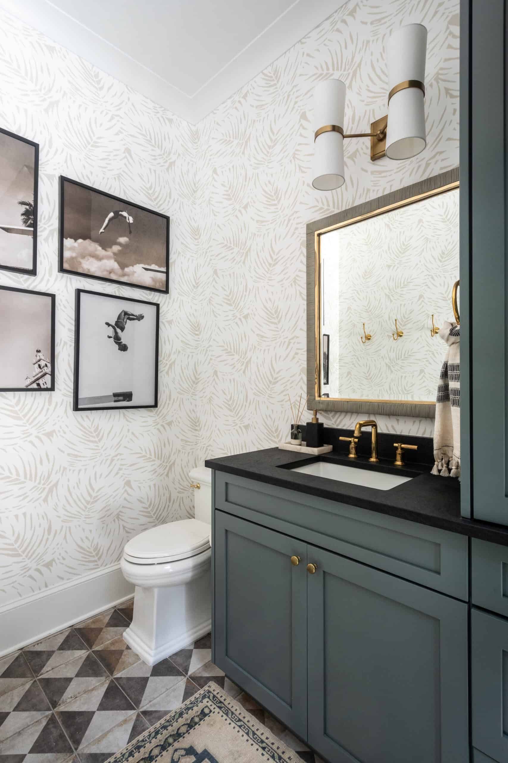 A powder room featuring a dark green vanity with a black countertop, gold fixtures, and geometric floor tiles. The room is decorated with a patterned wallpaper, black and white framed photographs, and a gold-framed mirror.