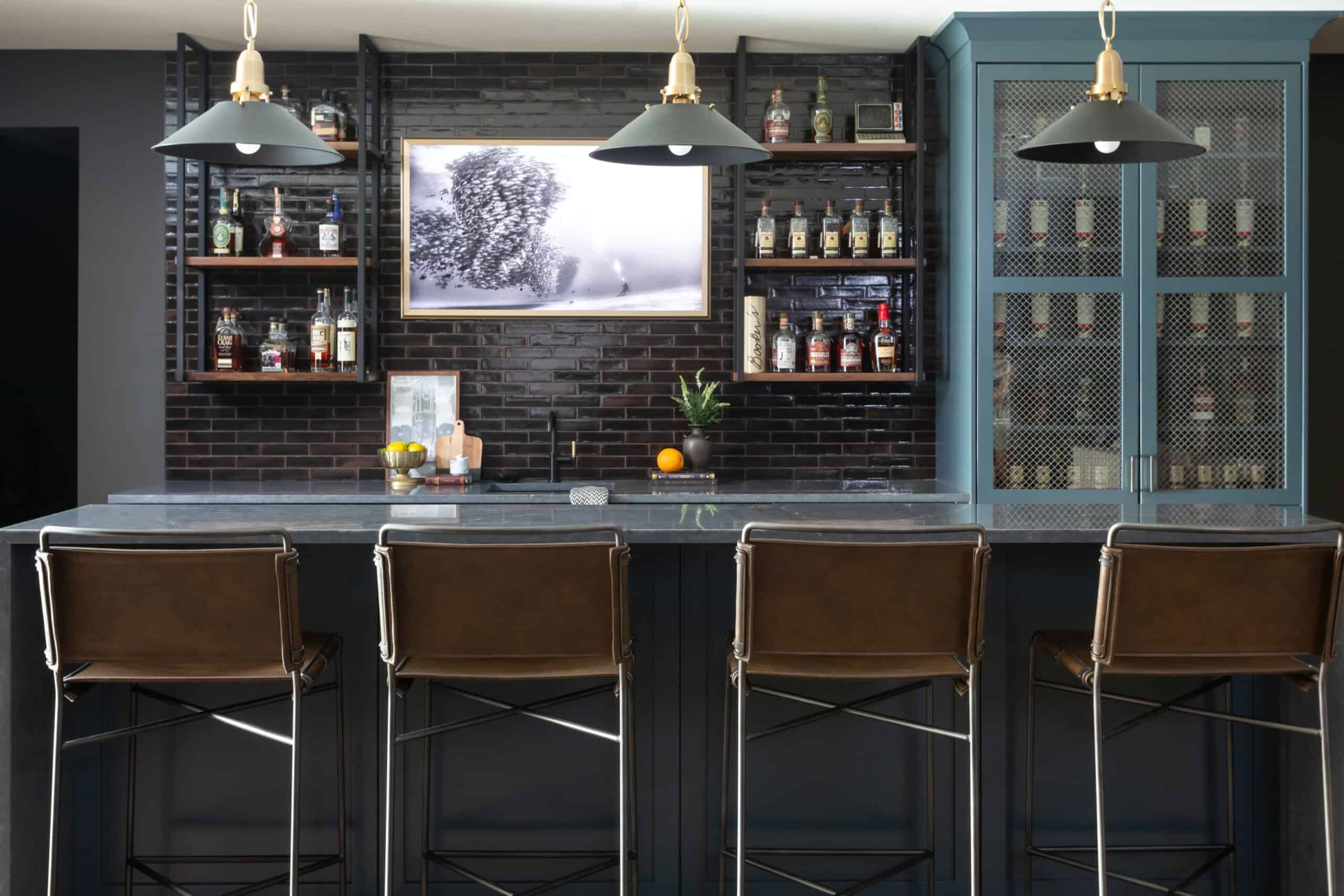 Stylish home bar with dark brick backsplash, black countertop, shelves with various bottles, and three brown leather bar stools. A framed black-and-white photo is mounted on the wall.