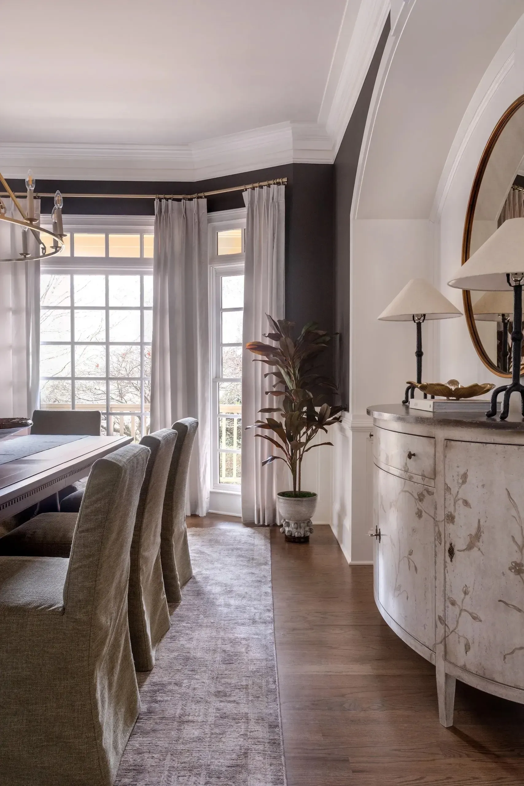 Floor-to-ceiling pale gray pinch-pleat curtains on a brass rod, framing a bay window in an elegant dining room with dark walls and white wainscoting.