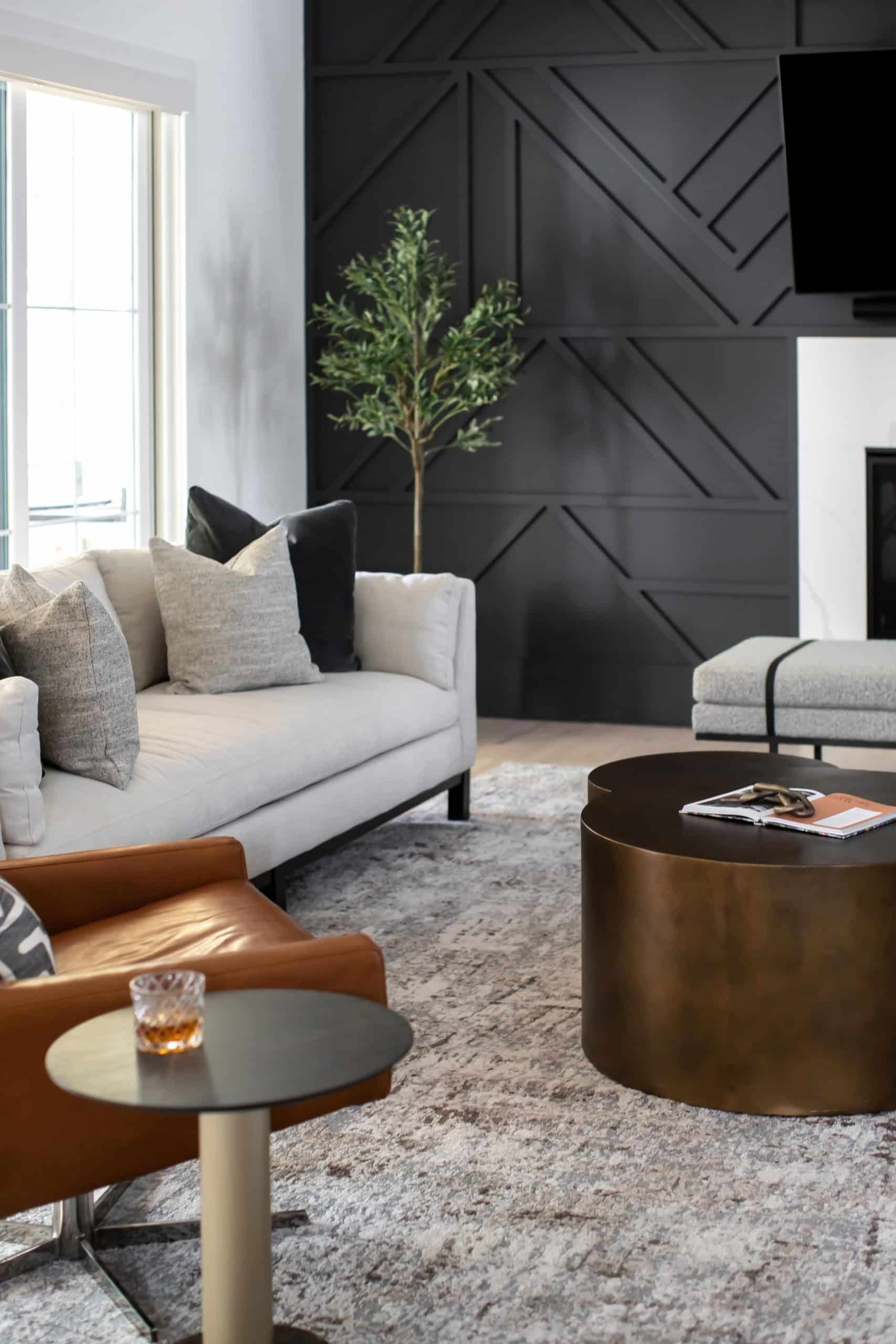 A modern living room with a white couch, black geometric accent wall, and a small potted tree. The room includes a round metallic coffee table, a leather armchair, and a small side table with a glass tumbler.