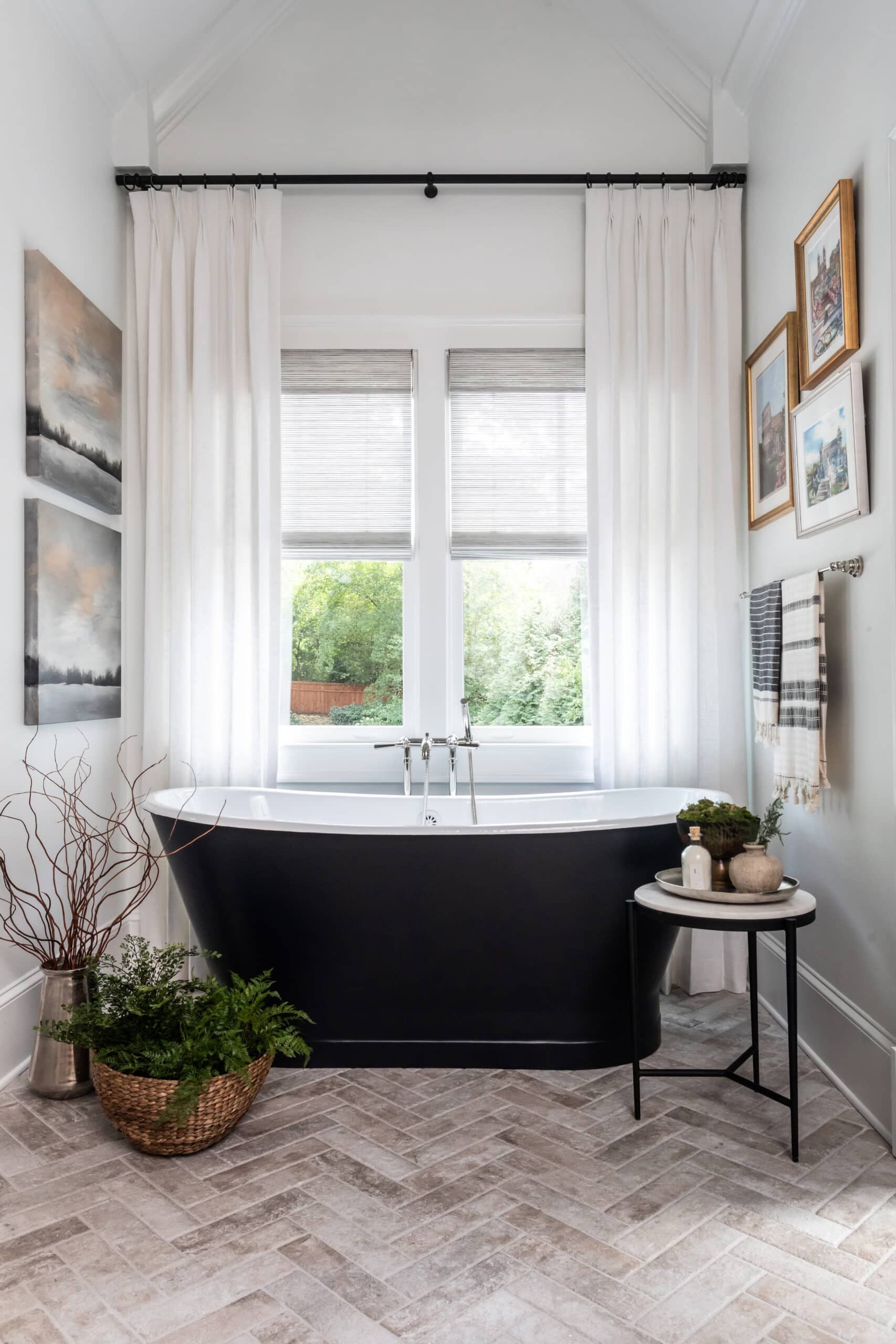 A bathroom with a black freestanding tub, white curtains, and a herringbone-patterned tile floor. The room is decorated with framed art, a wicker basket with plants, and a small side table with bath accessories.