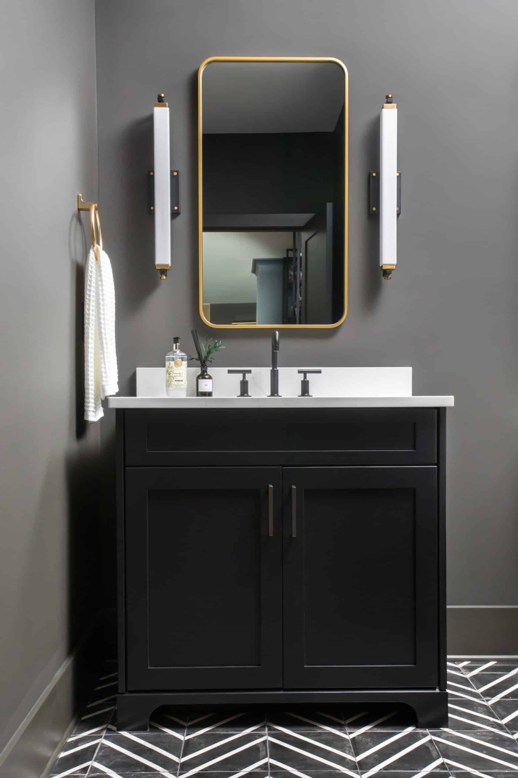 Elegant bathroom with a black vanity, white countertop, and dark gray walls. Features a rectangular mirror with a gold frame and modern lighting fixtures on either side.