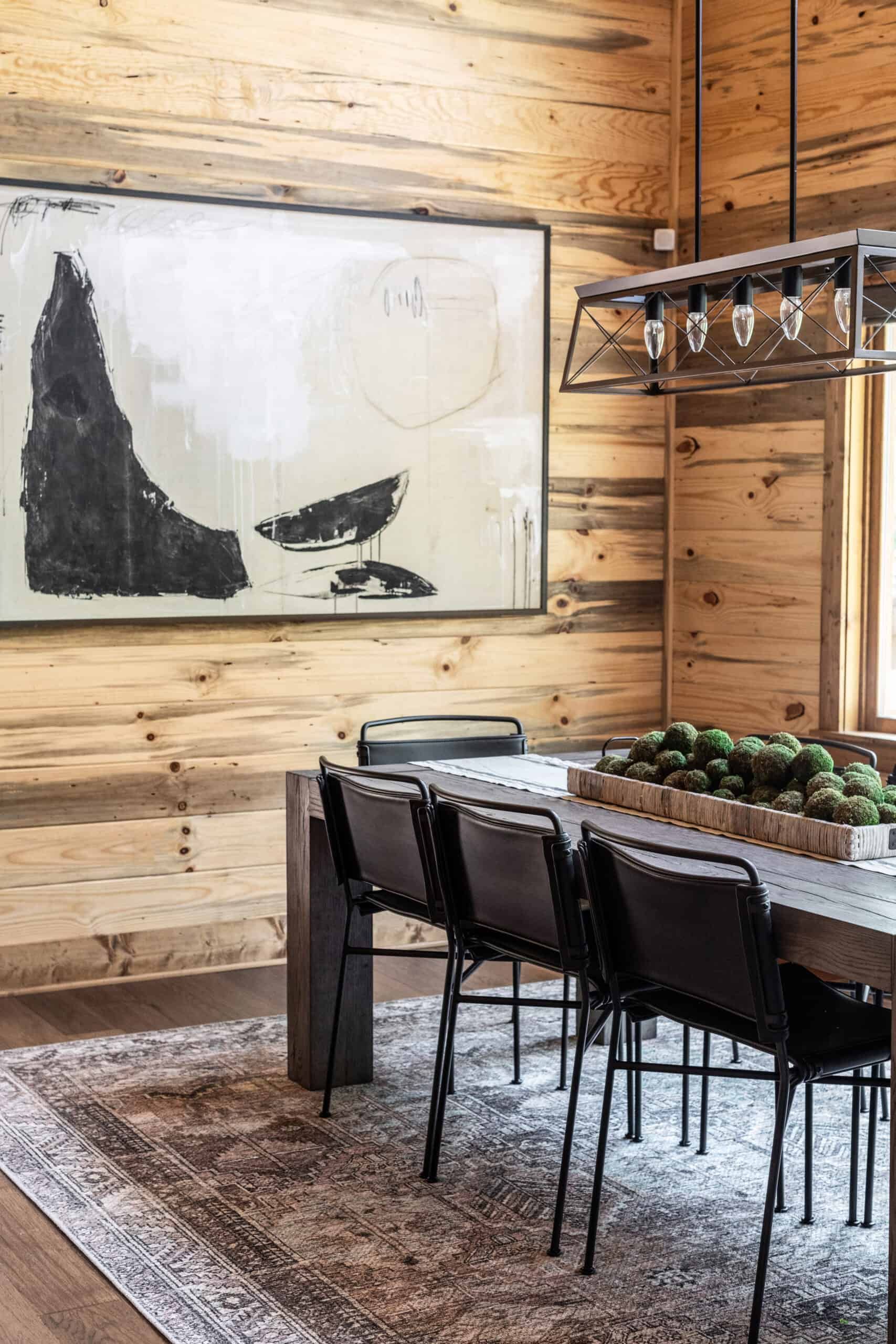 A rustic dining room with natural wood-paneled walls, a dark wood dining table, and black metal chairs. The room features a large black and white abstract painting, a modern chandelier, and a patterned area rug.