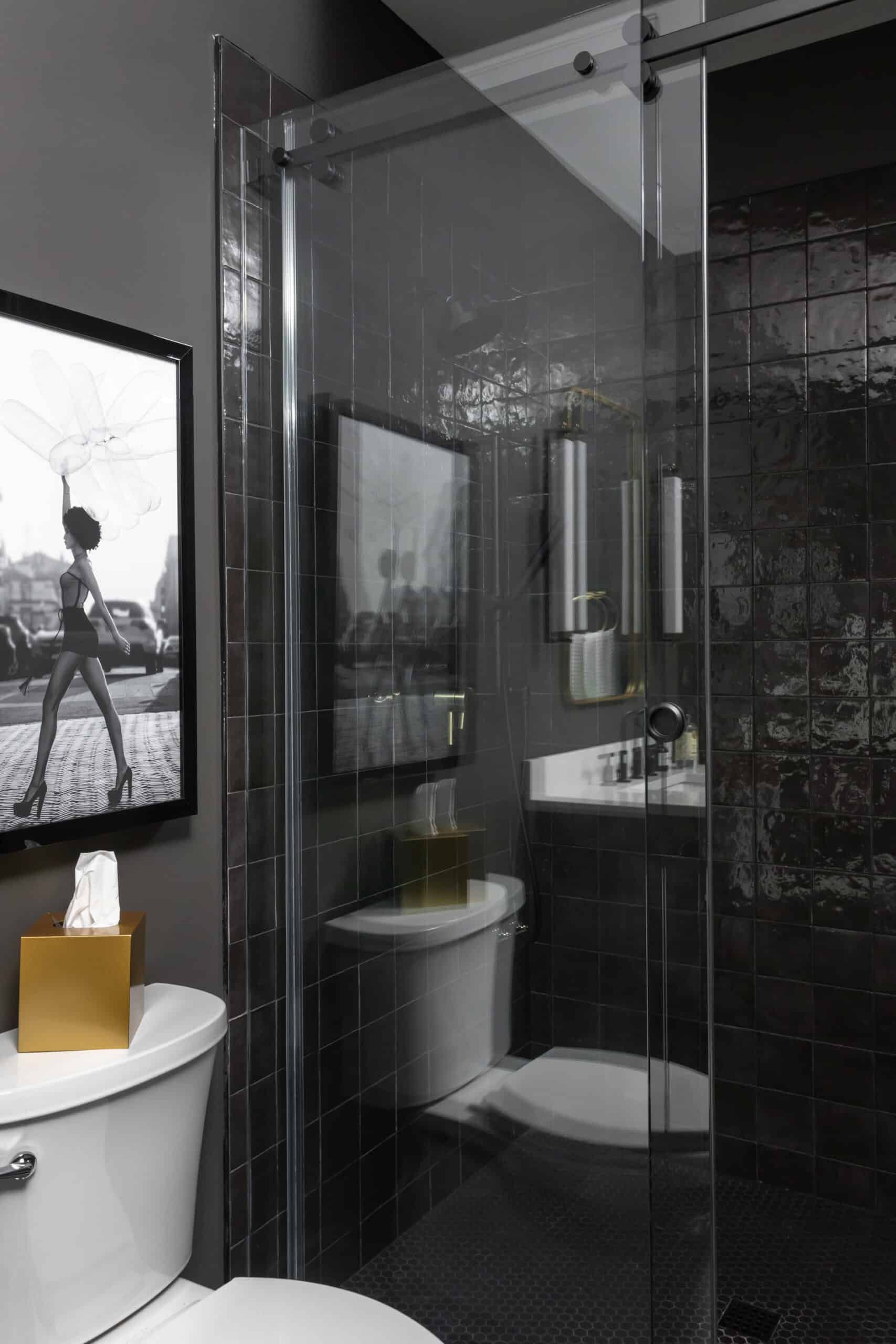 Modern bathroom with dark tiles and a glass shower door. A framed black-and-white photo of a model in heels adds a stylish touch. The space includes a white toilet and contemporary fixtures.