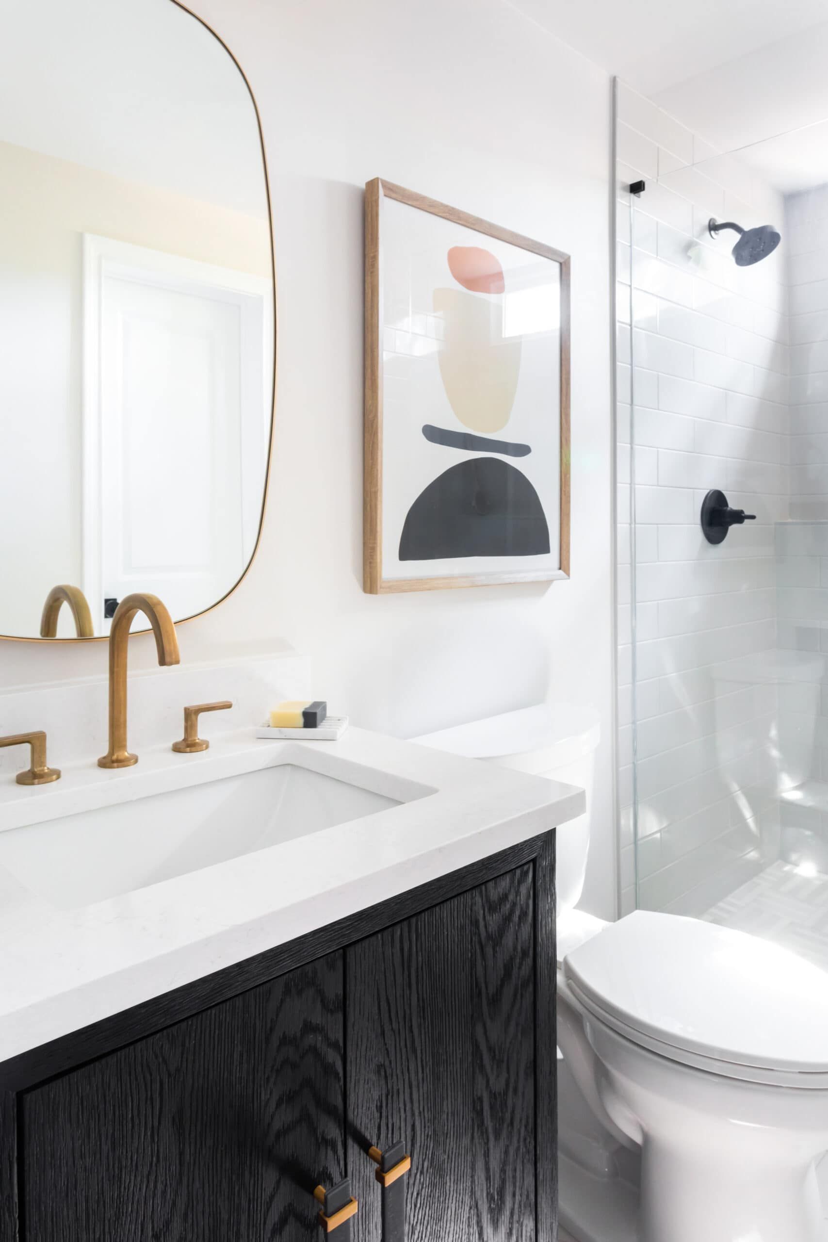 Contemporary bathroom with a white countertop, black vanity, and brass fixtures. Features a framed abstract artwork and a glass shower door with modern black fittings.