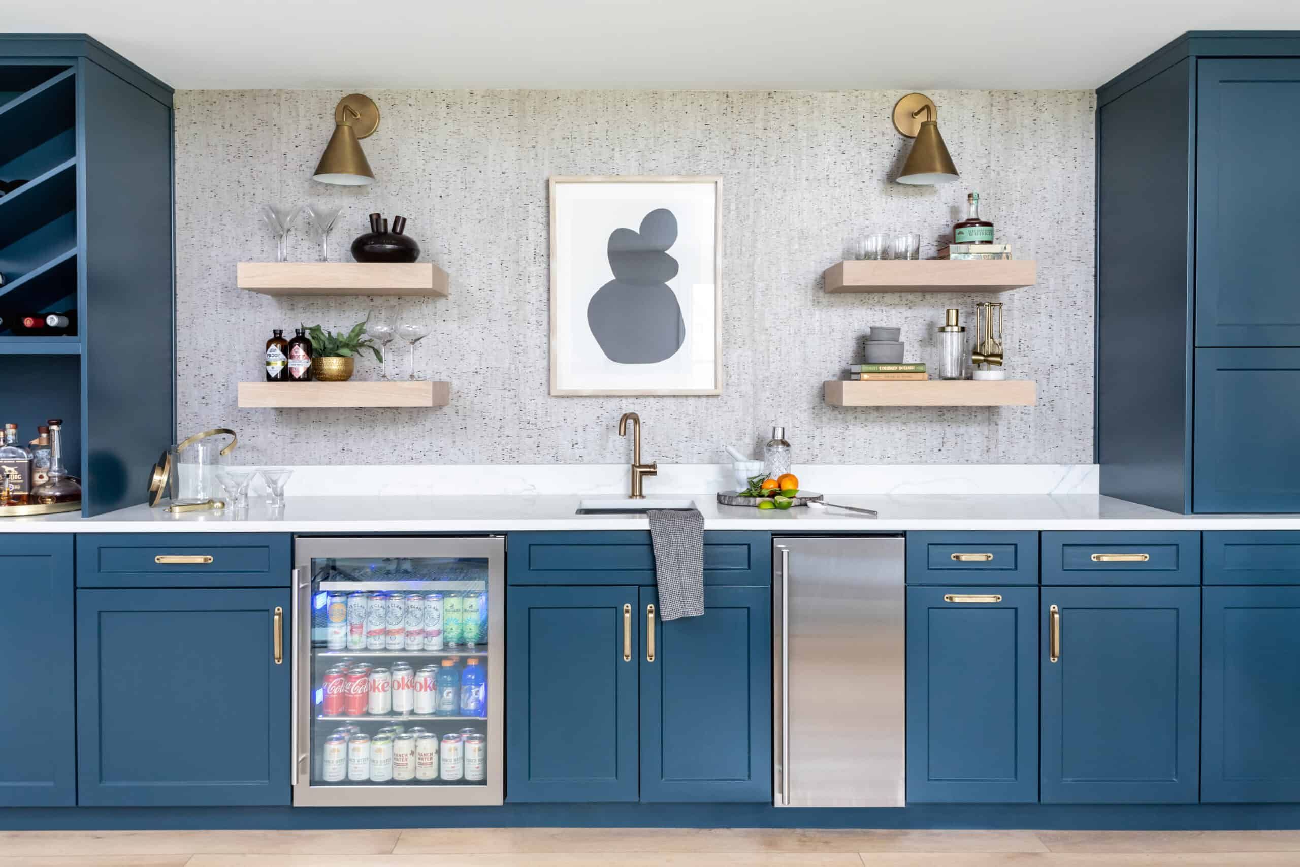 Bright and modern kitchenette with blue cabinets, white countertop, and wooden shelves. Features a mini-fridge with drinks, stainless steel sink, and contemporary decor.