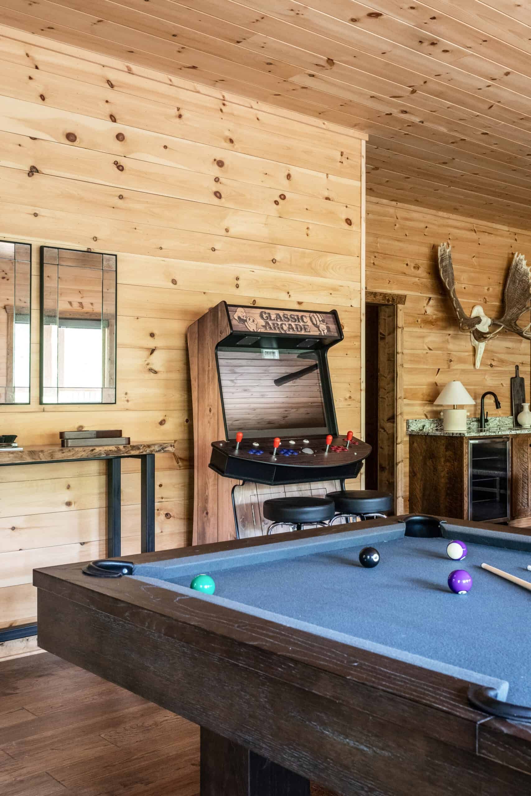 Rustic game room with wood panel walls and ceiling, featuring a classic arcade machine and a pool table. Mounted antlers and a kitchenette with granite countertops are visible in the background.