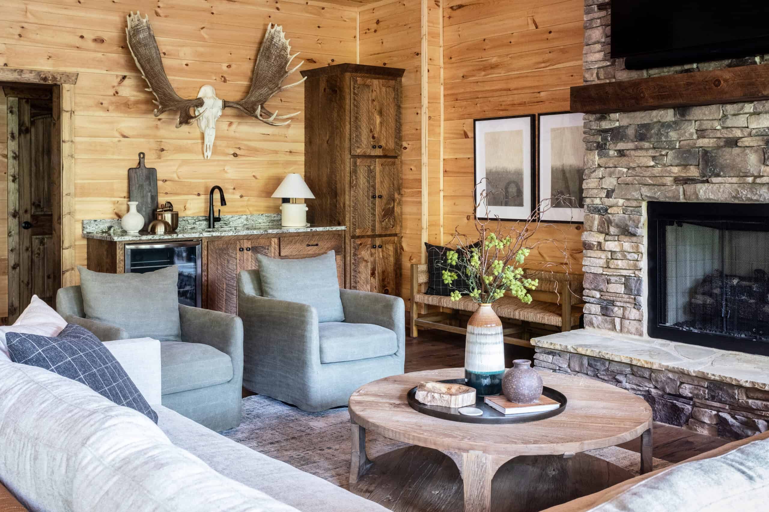 Rustic living room with wooden walls, a stone fireplace, gray armchairs, and a wooden coffee table. Decor includes mounted antlers and a small kitchenette with a granite countertop.