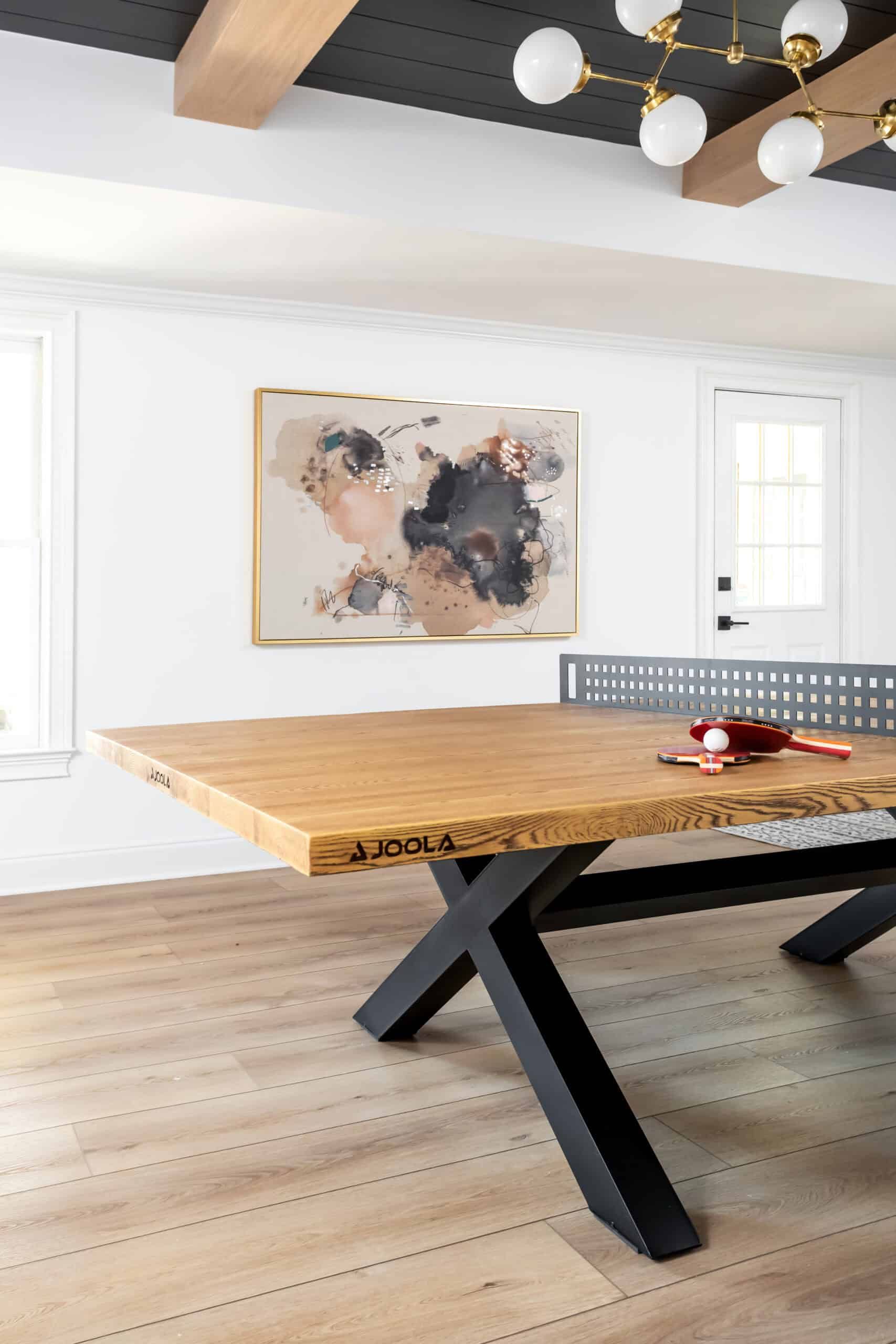 Modern game room with a ping pong table, featuring a wooden top and black legs. The room has a white wall with a framed abstract painting and a modern light fixture on the ceiling.