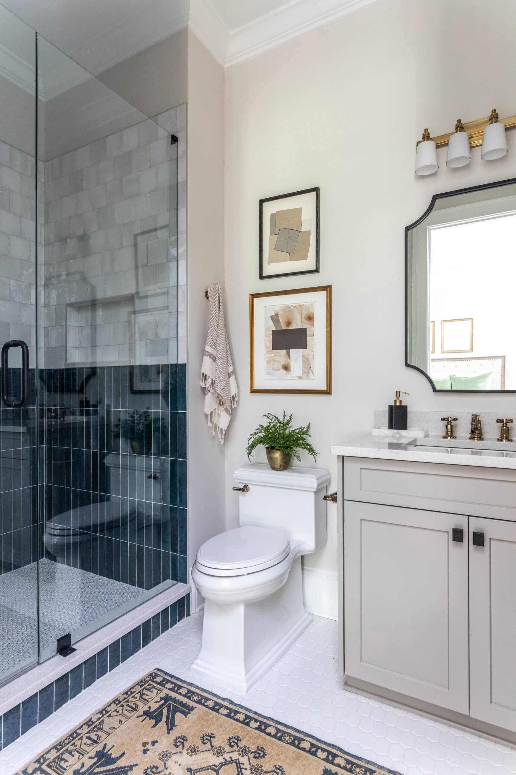 A bathroom featuring a glass-enclosed shower with dark blue tiles, a white toilet, and a gray vanity with a white countertop. The room is decorated with framed abstract art, a potted plant, and a beige and blue patterned rug.