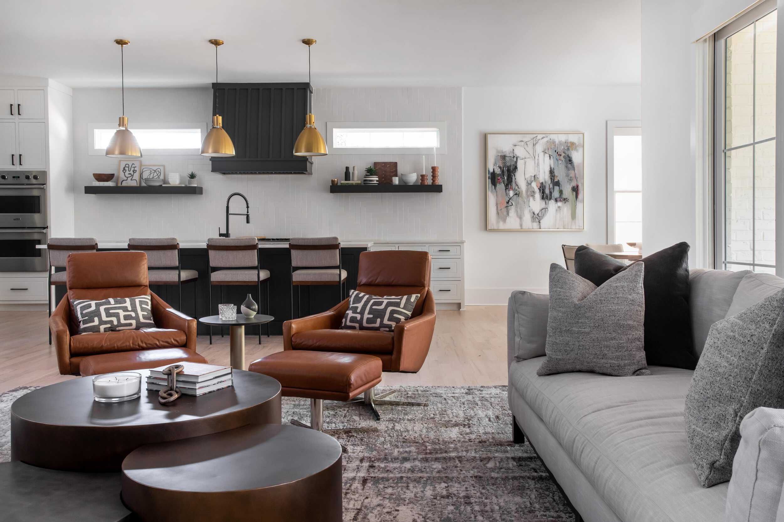 Two Eames-inspired chairs sit on either side of a minimalist, round end table with a three-tiered brown coffee table in front of them resembling tree trunks. A gray sofa sits to the right, with a kitchen island in the background underneath gold pendant lights