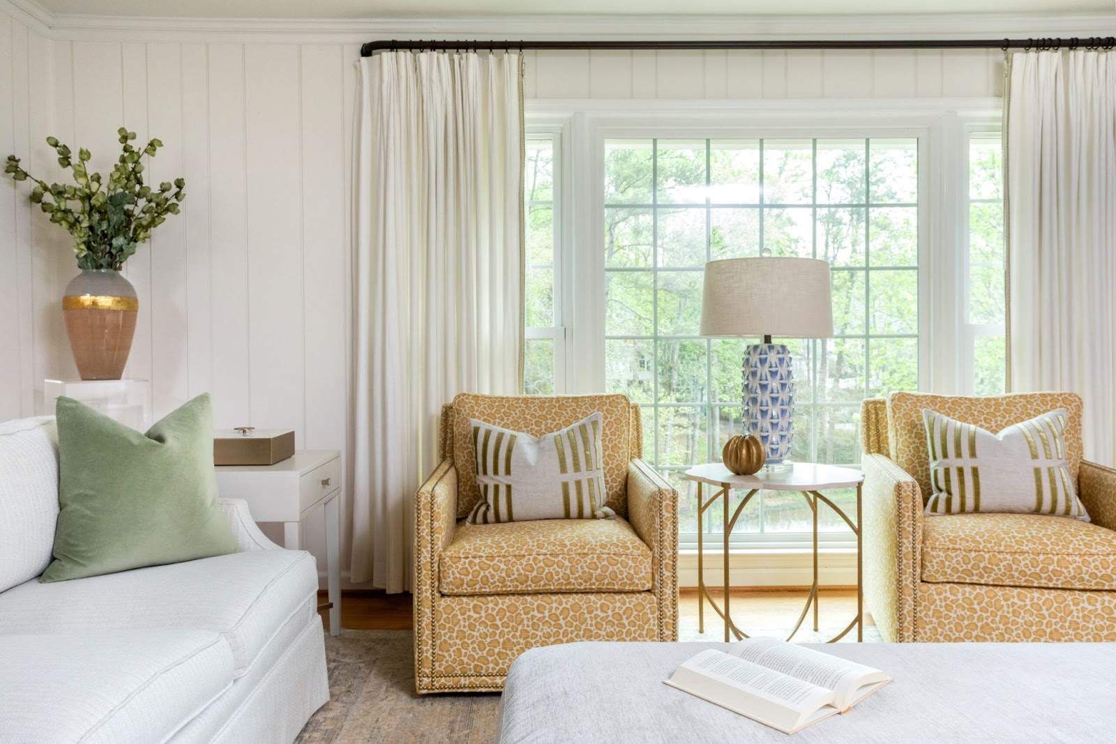 The corner of a white living room featuring two swivel cheetah print chairs, a white sofa and ottoman, green pillow, blue lamp, and gold vase with greenery. This is an example of a project where custom furniture design in Atlanta was offered.
