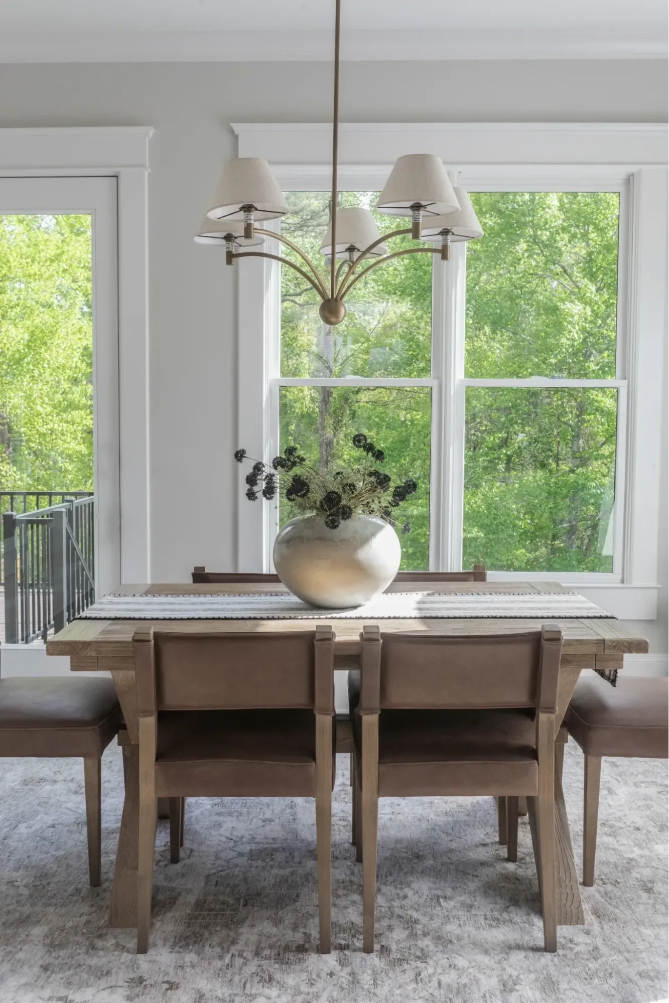 Modern dining room with an oak dining table, upholstered leather chairs, statement chandelier, and large windows overlooking wooded views.