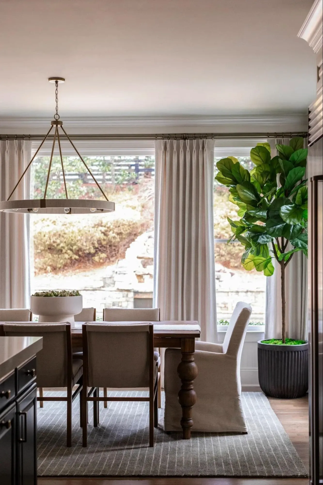 Elegant dining room with a wood dining table, custom drapery, statement chandelier, neutral upholstered seating, and large picture windows.