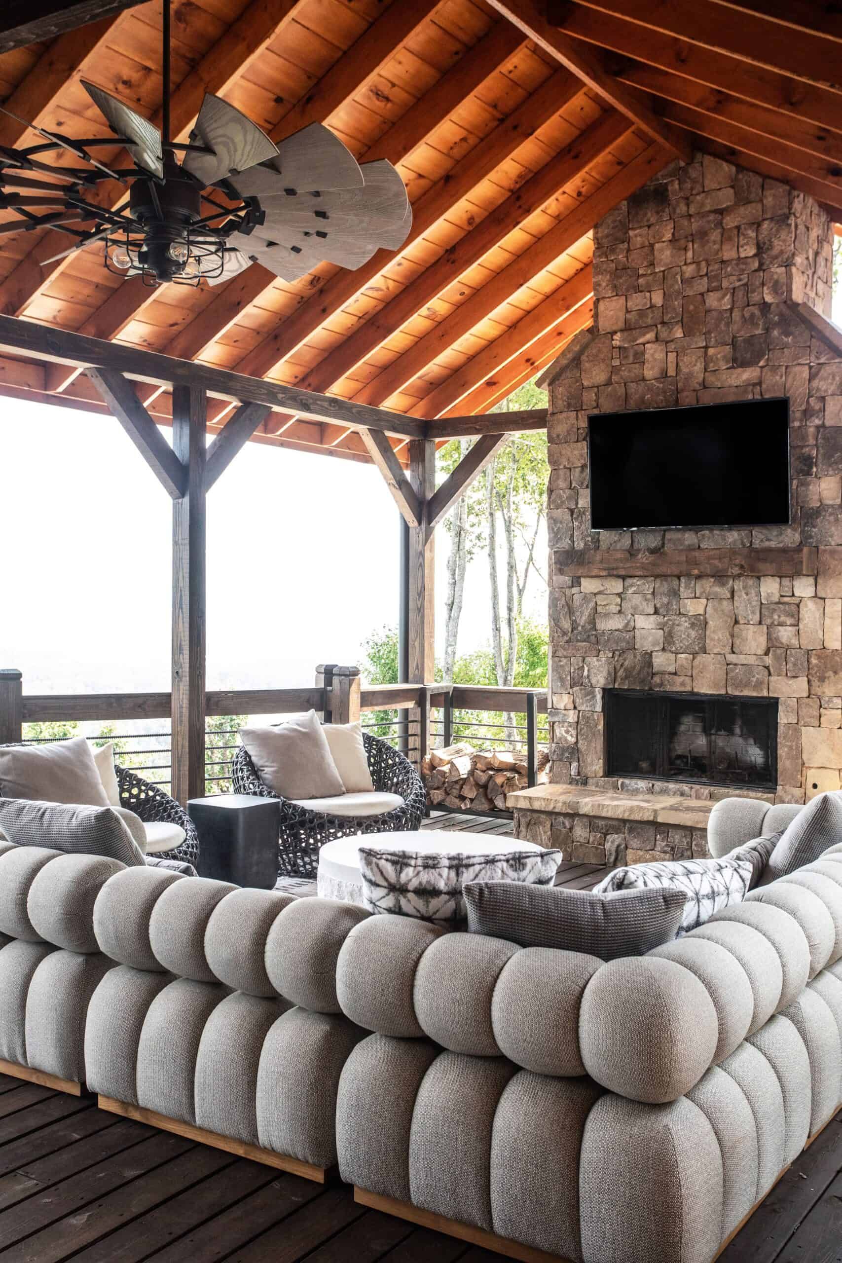 Outdoor balcony area with a gray sectional couch, Tv mounted on a stone fireplace, black metal and wood fan, with a cathedral roof.