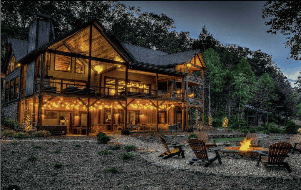 Exterior view of Whispering Water Lodge at night. In front of cabin is outdoor fireplace with wood chairs around it and glowing lights illuminate the impressive wood structure.