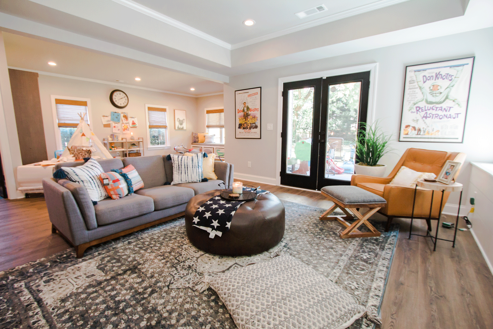 Modern living room with a gray sectional sofa, leather ottoman, and armchairs. Natural light streams through large glass doors. Decor includes framed posters, a clock, and a children's play area with a teepee.